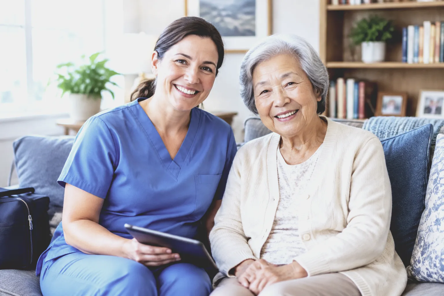 A smiling nurse in blue scrubs, holding a tablet, sits beside an older woman on her sofa
