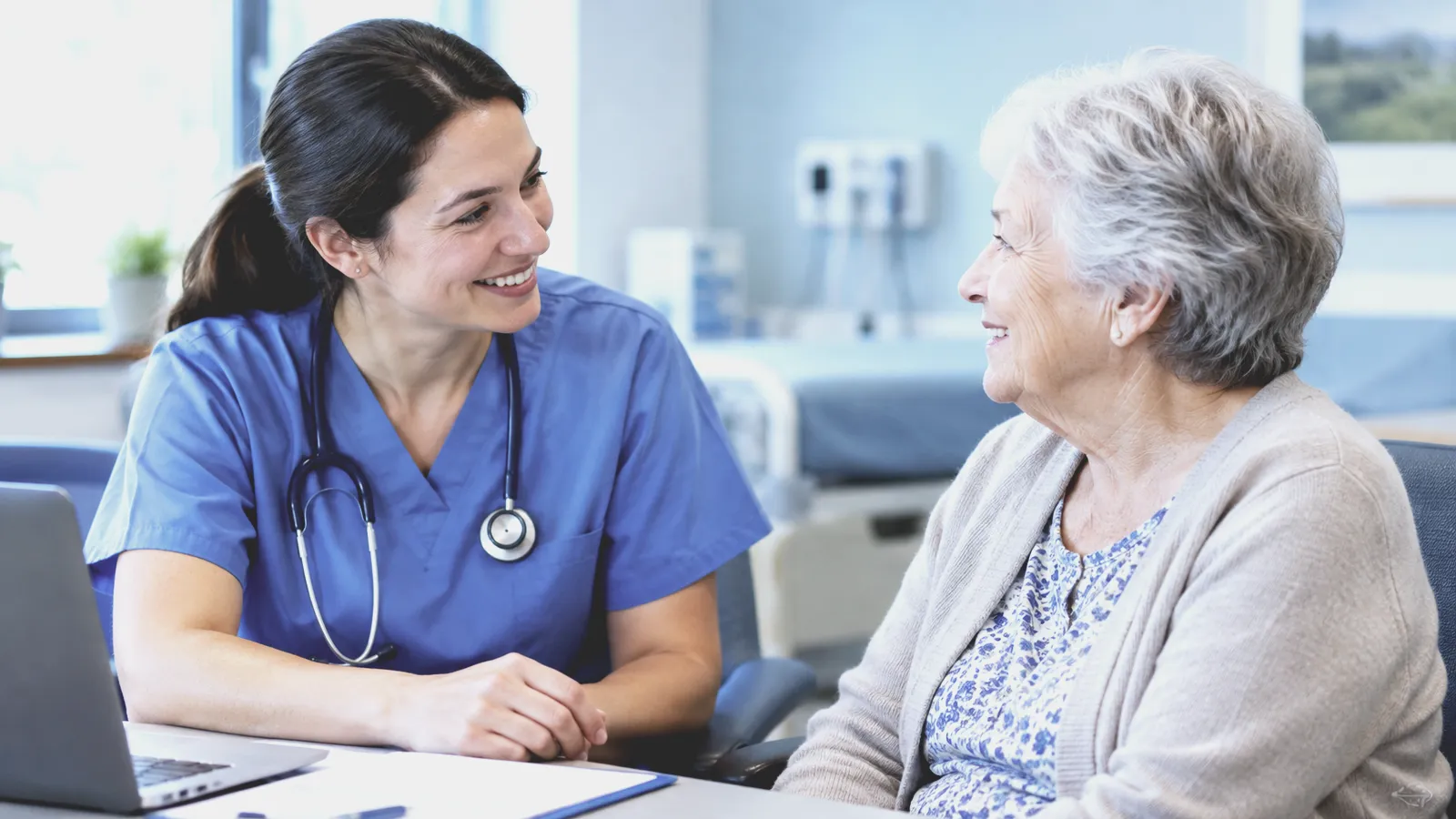 A smiling nurse in blue scrubs talks with an older woman across a desk