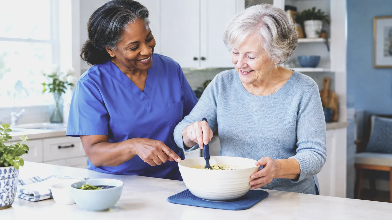 A therapist in blue scrubs points into a mixing bowl as an older woman stirs it at her kitchen counter
